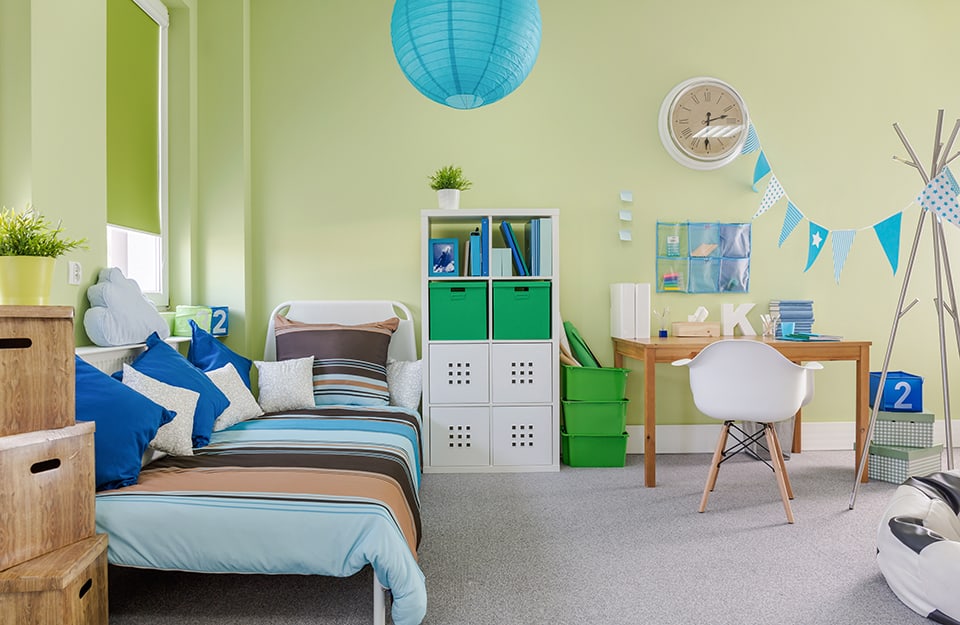 A bedroom with soft green walls, a white bed and striped linen in light blue and brown. On the bed are many pillows. Next to the bed is a bookcase with plastic and fabric books and drawers, next to green boxes and a wooden desk with a white designer chair. Above the desk is a large wall clock and a string with light blue triangular flags, reaching up to a coat rack. A spherical blue rice paper chandelier descends from the ceiling;