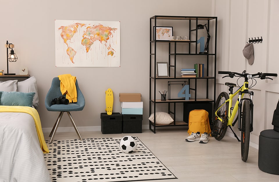 A little boy's bedroom with grey walls with a map of the world hanging on them. Next to the bed is a white carpet with regular black polka dots, a chair that serves as a bedside table, boxes with a yellow skateboard on it, a metal bookcase with books and objects, a yellow backpack, white trainers, a neon yellow off-road bike and hanging on the wall on a coat rack is a baseball cap;