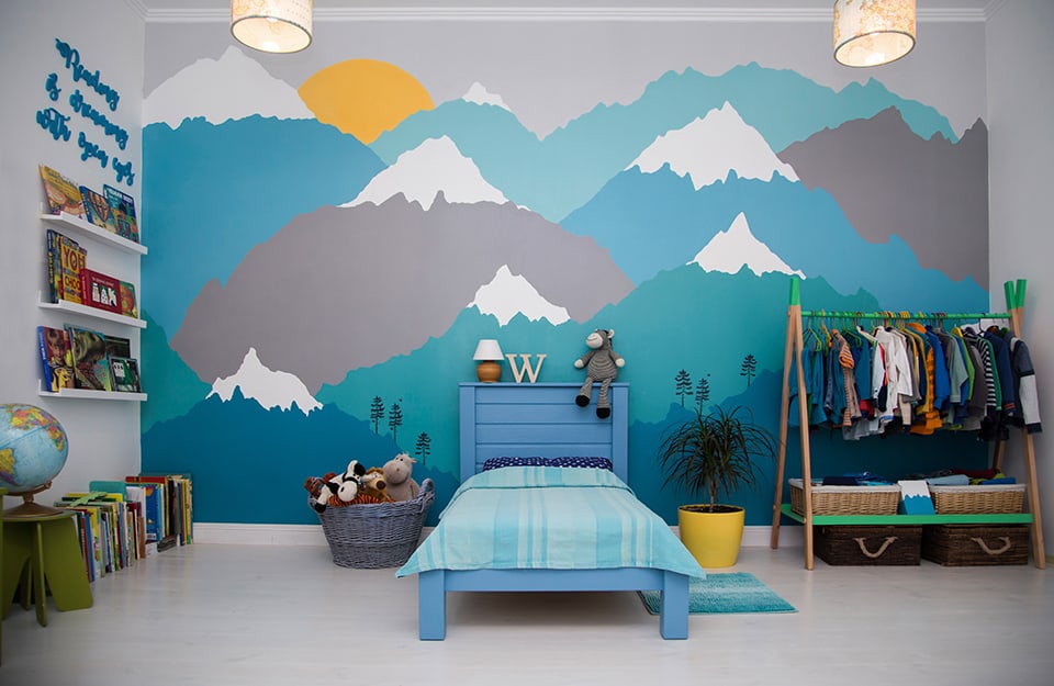 Bedroom with a large wall decorated with a mountain theme in shades of blue, grey and white. The bed is light blue and above the headboard there is an abat-jour, a wooden letter W and a toy monkey. On either side of the bed is a wicker basket full of toys and a plant in a yellow pot. To the right of the plant a clothes rack with clothes hanging from it. On the left wall shelves full of picture books and other books on the floor, next to a globe
