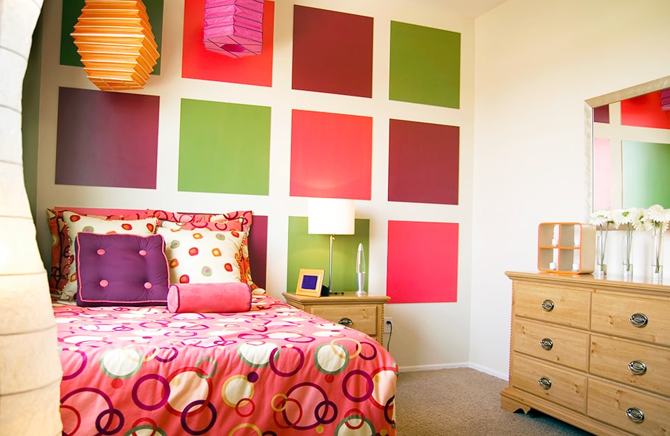 A multicoloured bedroom with a wall painted in squares of different colours (purple, green and magenta), a bed with a blanket with psychedelic 1960s motifs, multicoloured pillows, two Chinese-style chandeliers in orange and pink rice paper and a wooden dresser with knick-knacks and jars. Next to the bed is a wooden bedside table with a lava lamp on top;