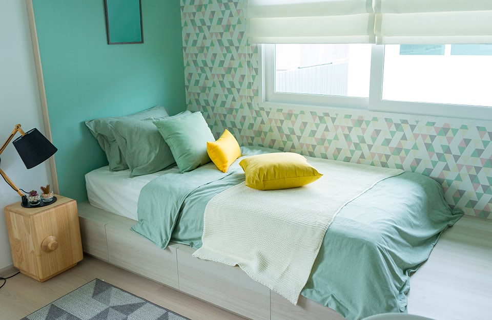 Corner of a boy's bedroom, with a bed with teal bed linen, teal wall behind the headboard and a wall with white and green wallpaper in geometric patterns. Next to the bed is a wooden bedside table with a reading lamp and a geometrically patterned rug;