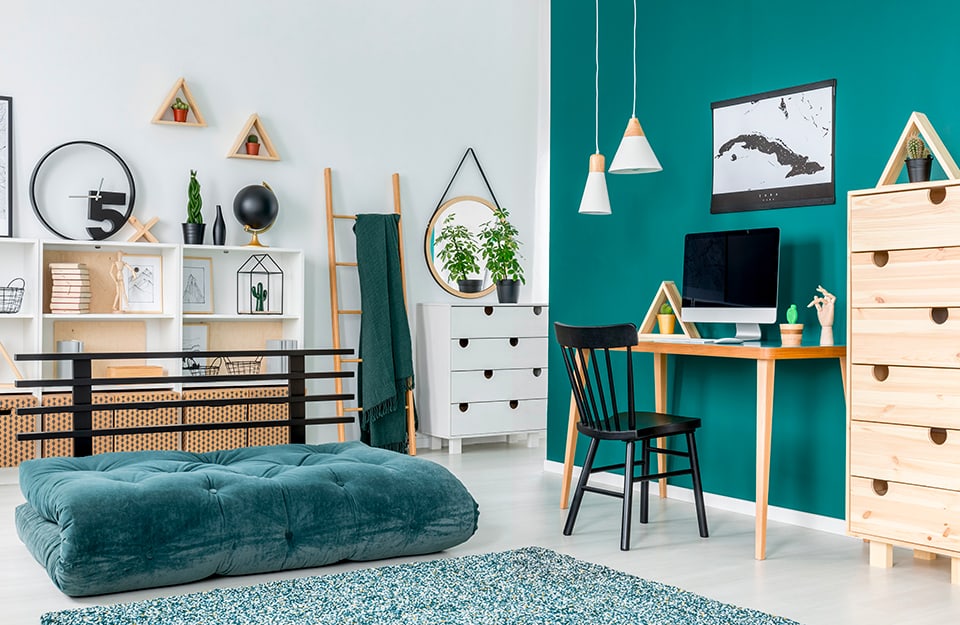 A teenage bedroom with a green wall, a fold-out sofa bed, white and wood-coloured bookshelves, many knick-knacks, a circular mirror hanging above a white chest of drawers and, in front of the green wall, a desk with a computer, a map of an island, conical chandeliers descending from the ceiling and a tall Scandinavian-style chest of drawers;