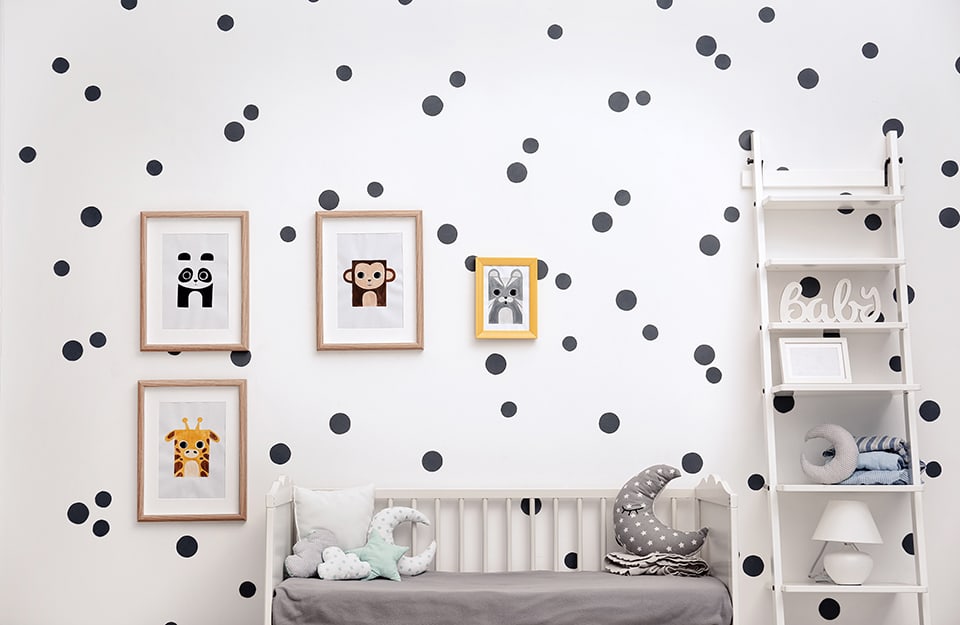 Paredes blancas de una habitación infantil decoradas con pegatinas de lunares grises de diferentes tamaños. También hay una cuna blanca sin barandilla, una estantería con escalera y láminas con animales estilizados