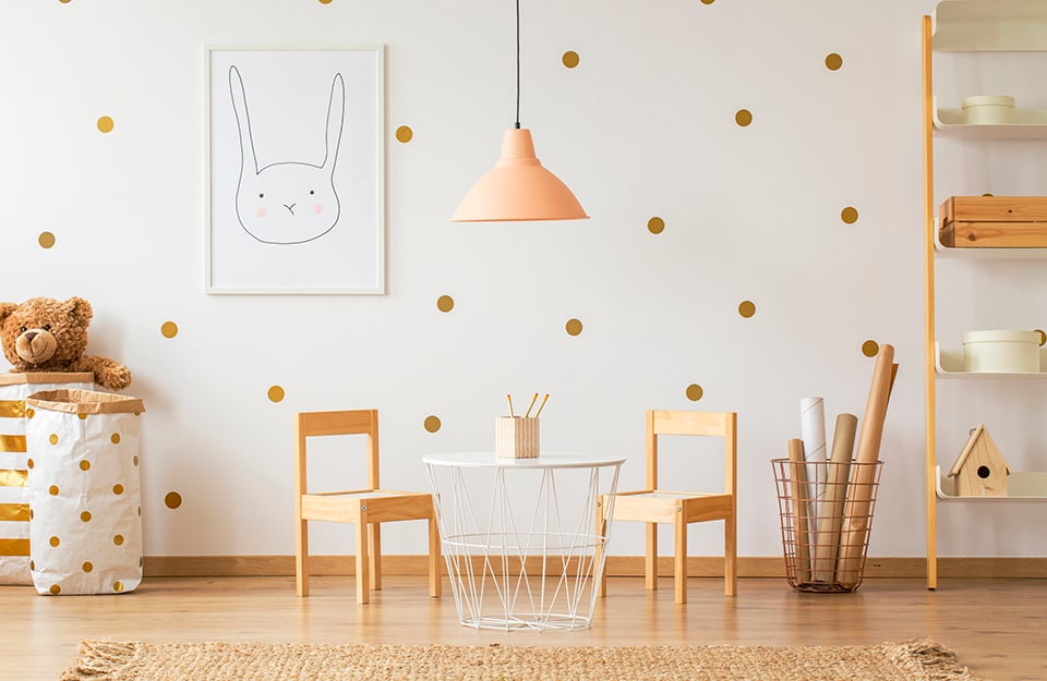 Children's room with a white wall decorated with metallic polka dot stickers. The room is Scandinavian in style, with wooden furniture (small chairs, ladder bookcase), a pastel pink chandelier, a print with a rabbit design, a white minimal metal table, a metal basket with cardboard rolls and paper baskets containing toys