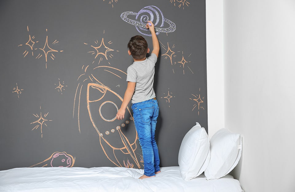 Niño de pie sobre la cama dibujando con tiza en la pared pintada con pintura de pizarra