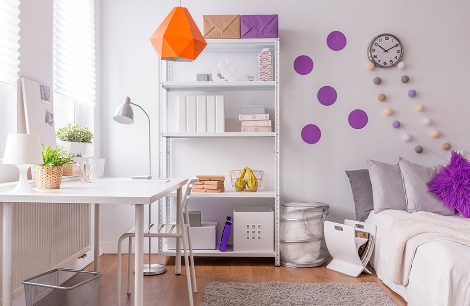 Little girl's bedroom with white walls decorated with purple polka dots, a colour that echoes the cushion on the bed and some other elements in the otherwise totally white room, with a desk, chair, metal shelf, magazine rack, bed and wall clock. The floor is parquet
