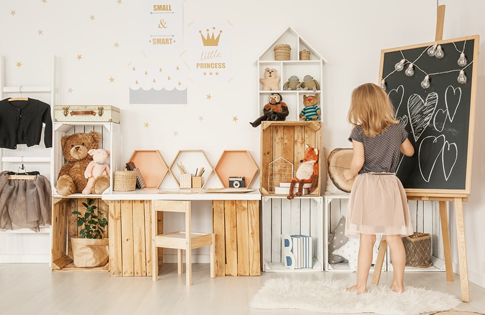 Blonde girl plays in her bedroom, furnished with recycled wooden fruit boxes, some painted white, and wooden storage boxes. There is a blackboard with hearts drawn on it. The wall is white with gold metal star stickers and prints hanging on it;