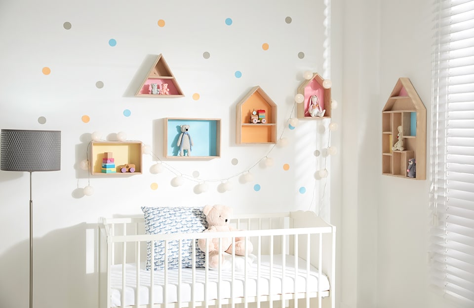 A baby's room with a white cot, white walls decorated with pastel-coloured polka dots and shelves in the shape of small houses and rectangles with pastel-coloured backgrounds containing toys. Next to the cot is a grey floor lamp and a window with white Venetian blinds can be glimpsed;