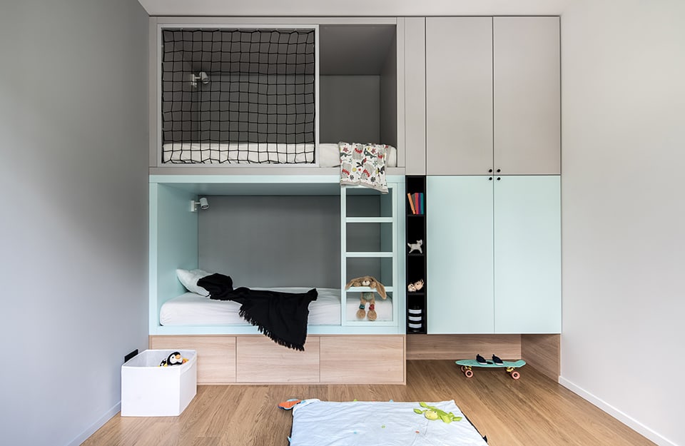 Children's bedroom in loft mode, with a structure that puts together a wardrobe and two beds, one on top of the other. The walls are grey and white and the wardrobe-bed is grey, pastel blue and natural wood. A skateboard and toys can also be seen;