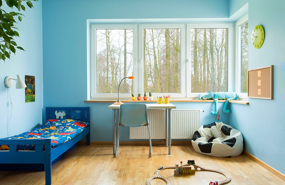Child's bedroom with a large corner window overlooking a tree-lined landscape. The walls are blue, the floor is parquet. The bed is blue, and in front of the window is a desk with a blue chair. On the desk are a lamp, some succulent plants and pencil cases. In the corner is a pouffe in the shape of a football and on the floor is a wooden toy car track;