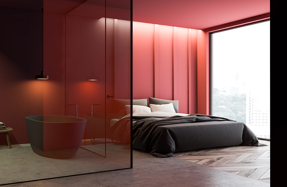 Una habitación con un gran ventanal que cubre toda una pared, con vistas a un paisaje urbano desde arriba. Las paredes de la habitación son rojas, al igual que el techo, y el suelo es en parte de parqué y en parte de hormigón. Paredes de cristal separan la cama del cuarto de baño, donde se ve una bañera oscura de estilo minimalista;