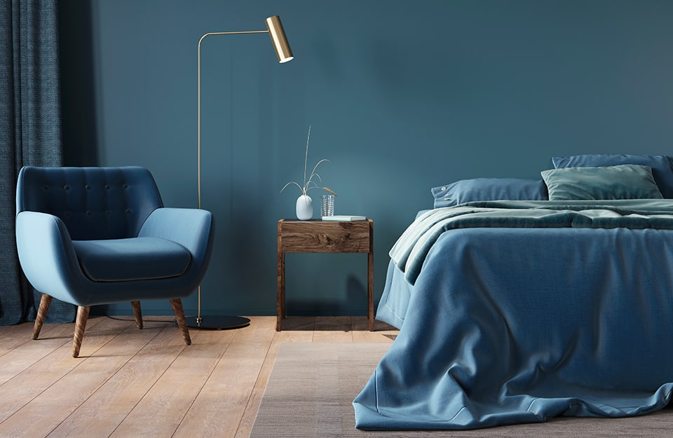 Bedroom all in shades of blue, with bed with blue linen, blue wall, blue Scandinavian-style armchair. The floor is a parquet with large boards. The bed is on a grey-beige carpet and next to the bed is a wooden bedside table with a book, a glass and a white jar on it. Between the armchair and the bedside table is a minimal, golden floor lamp;