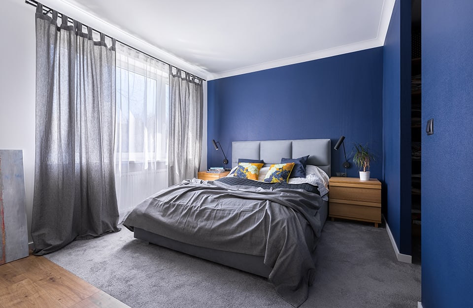 Bedroom in shades of blue and white. The bed is double and the headboard is upholstered. At the sides are two bedside tables in natural wood with essential lines and drawers. A white and grey curtain covers a long horizontal window and a radiator. The bed is on top of a carpet but part of the parquet floor can be glimpsed. To the right is an opening leading into another room;