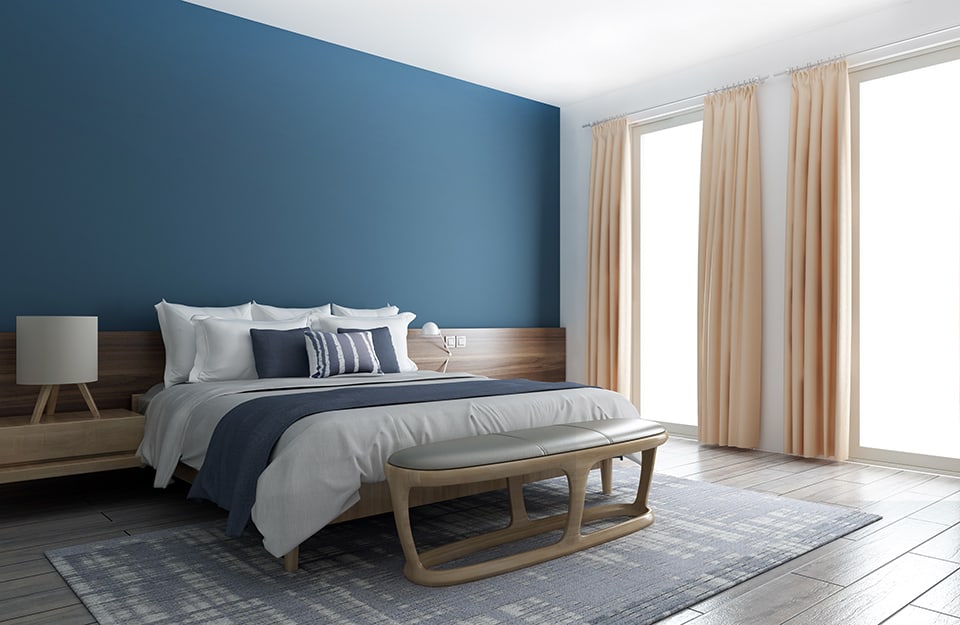 Large, bright bedroom in a sober modern style. The headboard of the bed is a long wooden band that covers the entire length of the wall and also incorporates the bedside tables. The rest of the wall is blue, while the ceiling and other walls are white. At the end of the bed is a large wooden bench with a soft seat and rounded lines. A large carpet in shades of blue and white is under the bed;