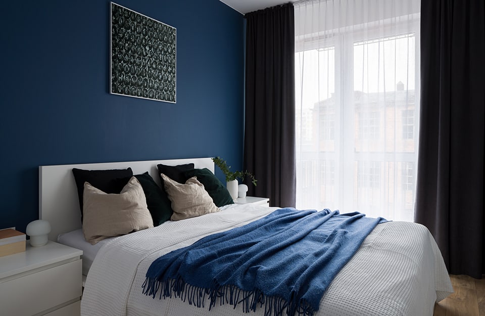 A sober, modern bedroom in a room with blue walls, white ceiling and parquet floor. A double French window, covered by a semi-transparent white curtain, opens onto a balcony. On the bed is a blue blanket and above the bed a dark abstract print. The comforters are white and plain, with drawers;