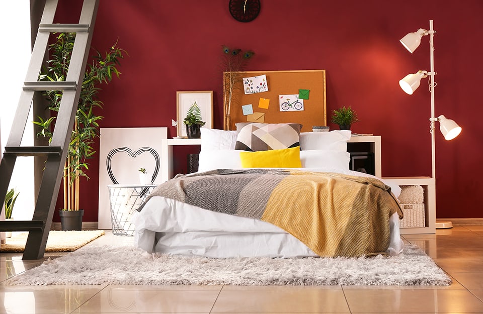 Open-plan bedroom with red walls. The bed is on top of a long-pile carpet and instead of the headboard there are horizontal shelves, above which are plants, prints and a cork board. Next to the bed, to the right is a floor lamp with three spotlights, to the left a small table, a painting and a potted plant, while a metal staircase leads to a mezzanine;