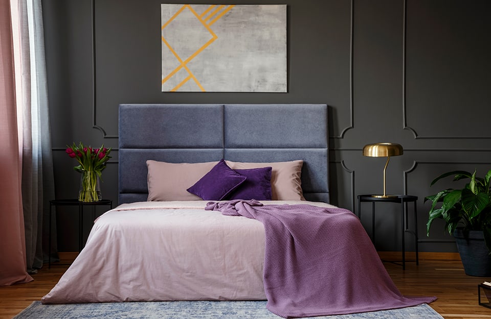 Elegant bedroom in shades of black and purple: black framed wall, double bed with purple headboard and pink and purple bed linen, carpet under the bed, parquet floor, two black metal side tables with a metal lamp on one and a vase with red tulips on the other. Above the bed is a grey and yellow abstract painting and to the right you can see part of a potted plant;