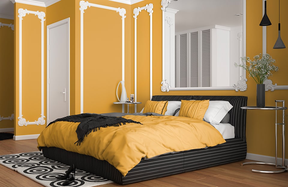 Large bedroom with yellow walls decorated with white baroque frames. Behind the bed is a large mirror with frames that echo those on the walls, and in which a white wardrobe can be seen reflected. The bed has a black frame and yellow and white bed linen. At the end is a white carpet with black circular geometric patterns. On either side of the bed are light metal coffee tables with vases on them. The floor is parquet