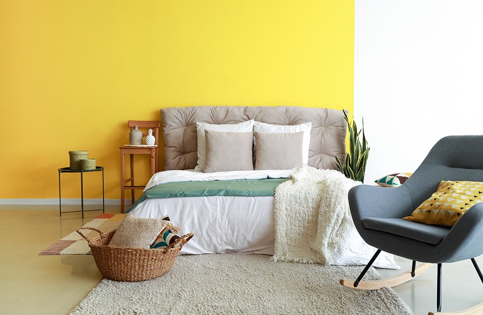 Bedroom with a two-coloured yellow and white wall (yellow on the left and white on the right). The double bed has a grey upholstered headboard. There are rugs at the sides and bottom of the bed, where there is also a wicker basket with cushions inside, and a blue Scandinavian-style rocking chair. At one side of the bed a plant can be glimpsed. At the other a high stool with a backrest, used as a bedside table, and a small black metal table with two vases on it. The floor is light