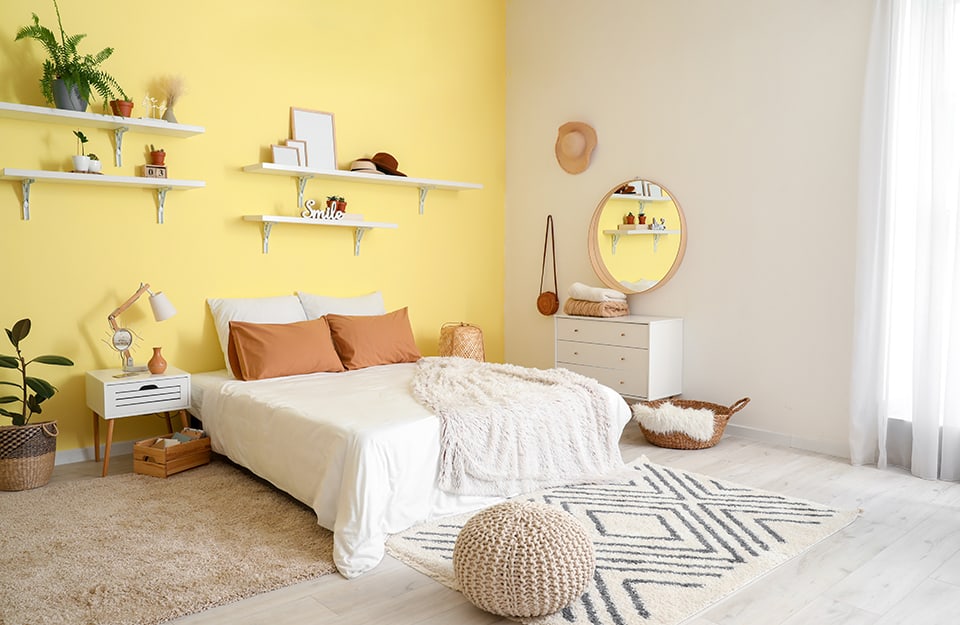 Boho-chic style bedroom in shades of yellow and white. The main wall, behind the bed, is pastel yellow, with several shelves holding knick-knacks, seedlings and frames. The bed is double, with a woven wooden lamp on one side and a Scandinavian-style wooden bedside table with a drawer and lamp on the other. At the end and to one side of the bed are light-coloured carpets. On the floor is a white parquet floor. On the white side wall hangs a circular wooden mirror, a straw hat and a vintage leather handbag, and a white chest of drawers. On the floor a wicker basket and a pouff