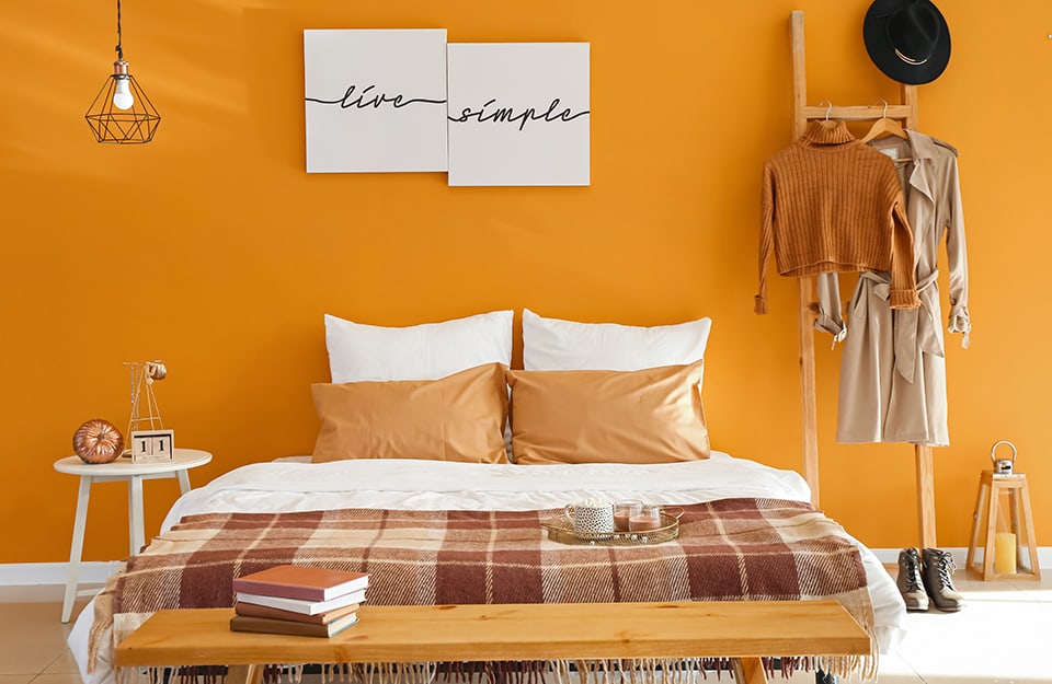 Bedroom in shades of orange. The wall is orange, with two canvases hanging with 'live simple' written in cursive. On a ladder leaning against the wall hang clothes (jumper and mackintosh) and a hat. At the foot of the ladder are a pair of shoes and a wooden lantern with a yellow candle inside. The bed has white and orange bed linen and a checkered blanket in shades of brown. At the end of the bed is a simple wooden bench with books on it. At the side of the bed is a small white wooden table with knick-knacks. A small black metal chandelier descends from the ceiling;