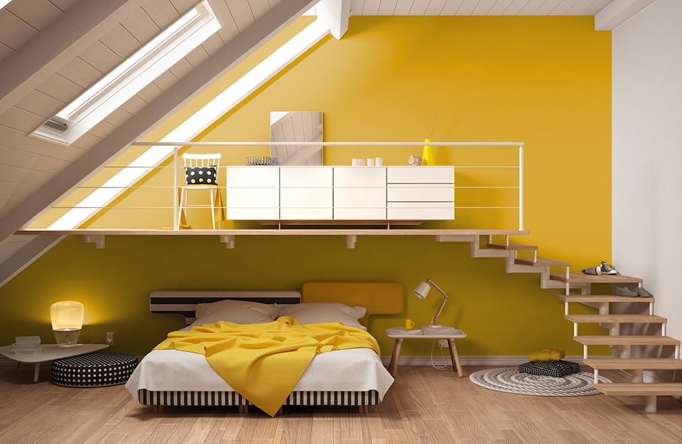 Open loft space with exposed floorboards, with a bed under the loft. The bed has a white blanket, and there are small tables at the sides. The headboard of the bed is asymmetrical, consisting of two different sized elements, one with black and white horizontal stripes and the other yellow. From the parquet floor, via open wooden stairs, one goes to a small mezzanine, where there is a white chest of drawers, a mirror and a white chair. The main wall (the one behind the bed and the loft) is yellow while the others are white;