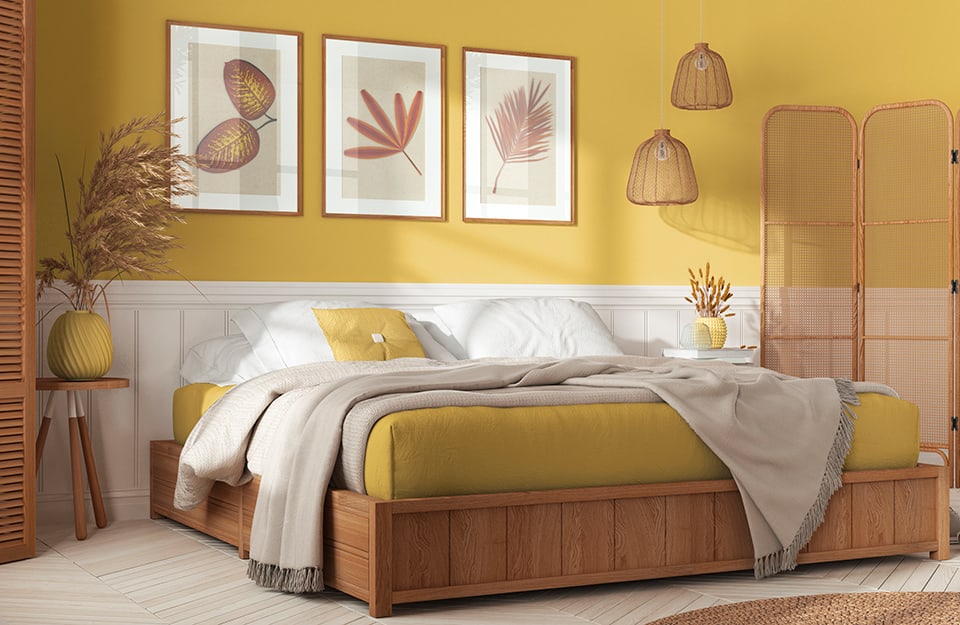 A bedroom in shades of yellow, with a double bed with a wooden frame and yellow and white bed linen. On either side of the bed are small three-legged tables with yellow vases on top. A wooden screen also appears on one side and two wicker chandeliers descend from the ceiling. On the yellow wall behind the bed is a low white paneling and above the bed some botanical-themed prints. The floor is light parquet;