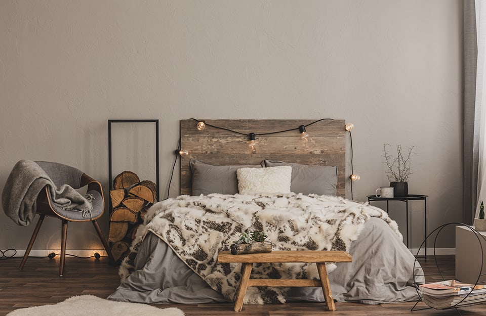 Bedroom in shades of grey and natural wood. The floor is dark parquet. The wall is grey. The bed has a headboard of reclaimed vintage wood, with a string of LED lamps resting on it. The bedding is white and grey and there is a long-haired blanket that looks like fur. At the end of the bed is a vintage, rustic-style bench. On one side of the bed is a black metal coffee table with a white enamelled cup and a black jar on top. On the other side of the bed is a rectangular metal frame with fireplace wood inside. Next to it is a Scandinavian-style armchair with a grey seat. A white carpet and an essential metal magazine rack can be seen on the floor;