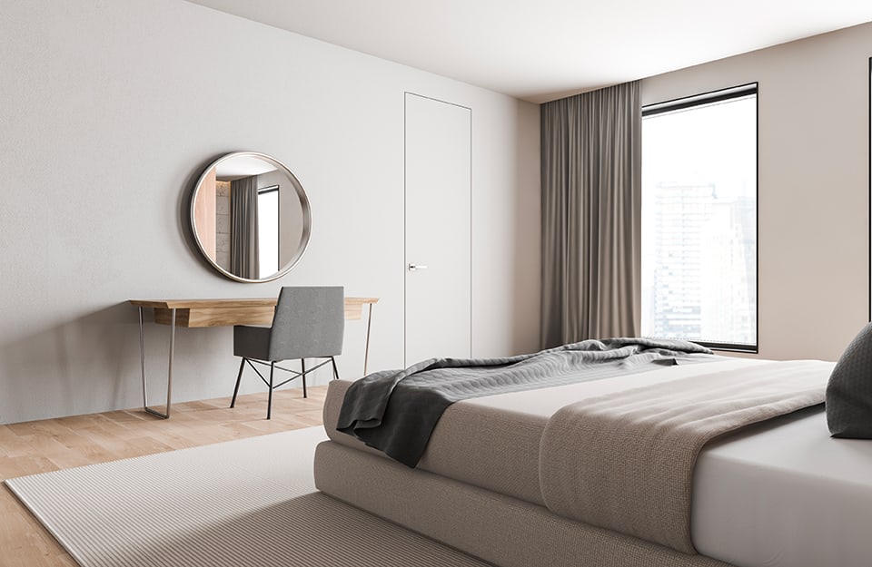 Spacious and bright bedroom in minimalist style, seen from the corner where the bedside table is. The walls and ceiling are a very light grey and the shape of a door can be seen on the back wall. There is also a desk with chairs and, above it, a circular mirror with a wooden frame. From the window, partially covered with a grey curtain, one can see an urban panorama. The bed has a grey upholstered frame and sits on top of a grey carpet as well. The floor is parquet