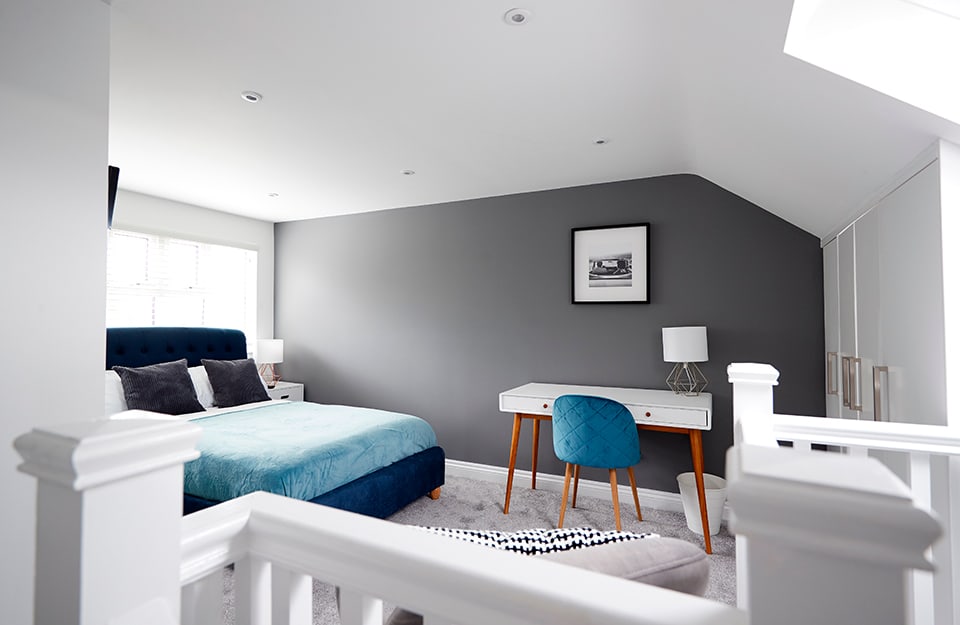 Attic bedroom in shades of white and grey. We see it from the point of view of someone coming up the stairs. The bed has a dark blue upholstered frame and bed linen in white, light blue and grey. The wall next to the bed is dark grey and there is a white modern style desk, a blue quilted chair and, hanging on the wall, a black and white photograph. There is carpet on the floor and at the end of the room, on the opposite side of the bed, a white built-in wardrobe. The rest of the walls are white and there is a skylight and a window behind the bed;