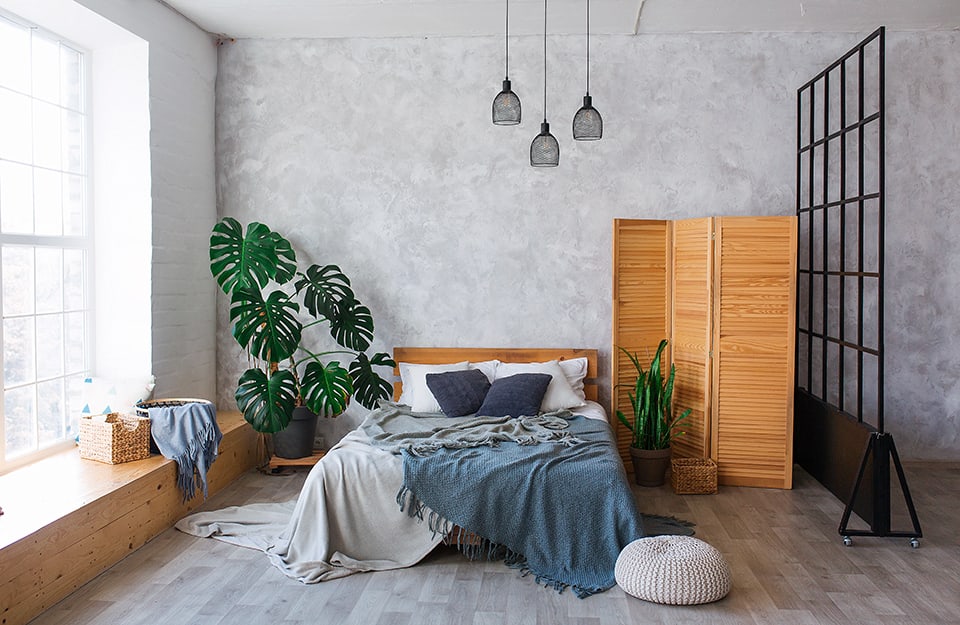 Bright bedroom with grey parquet floor and light grey sand-effect back wall. Under the window a long natural wooden frame serves as a bench. The double bed has a wooden frame and headboard. On one side is a large plant (monstera) in a pot. On the other is a wooden screen and a small potted plant. A black metal partition divides the space into what appears to be an open space;