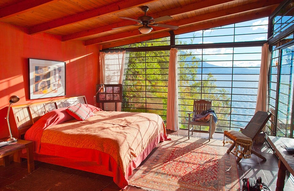 Bedroom in an attic with beamed ceiling and large windows overlooking the sea. The walls are red. The floor is terracotta, with several carpets. Above the bed there is a painting and in the room there is vintage furniture and several seats;