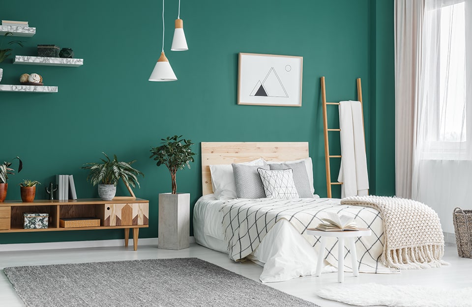 Boho chic style bedroom with white parquet floor and deep green main wall. The bed has white bed linen and a light natural wood headboard. To the right is a wooden ladder used as a coat rack, and there is a window covered by a semi-transparent white curtain. At the end of the bed is a small white wooden table with an open book on it, and a white carpet. Above the bed, a print with a mountain in minimal geometric black and white style. Two similar but differently shaped white conical chandeliers descend from the ceiling. To the left of the bed is a concrete parallelepiped shaped vase with a plant inside, and a horizontal wooden shelf in modernist style. Above, fixed to the wall, three white marble-effect shelves with knick-knacks