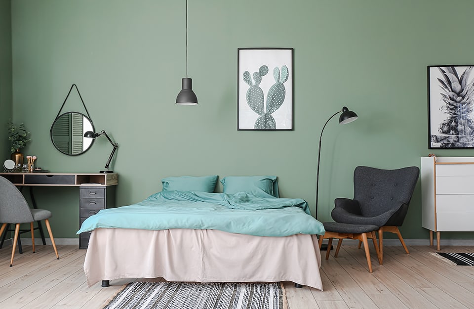 Large bedroom with green walls and light parquet floor. The bed is double, with grey and teal linen. Above the bed a botanical print. On one side of the bed, a black metal floor lamp, a grey Scandinavian-style armchair with matching, separate footstool, and a white cabinet with drawers with a black and white botanical-themed photo on top. On the other side of the bed, an essential dressing table with drawers, in wood and black, with a chair and, hanging on the wall, a circular mirror with a black frame;