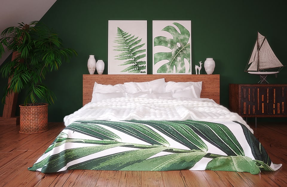 Attic room with bedroom. The floor is parquet, the back wall is green. The bed has a wooden frame, with a large headboard, above which are white vases and two botanical-themed prints. The blanket is also botanically themed. On either side of the bed is a large plant on one side and on the other an ethnic-style wooden cabinet with a model of a sailing boat on top;