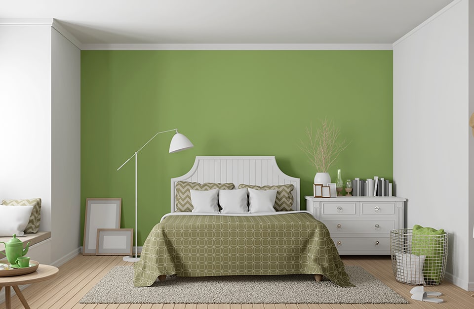 Modern bedroom in vintage style, in shades of white and green. The wall behind the bed is green (the others are white) and the bed is in white painted wood with a headboard with rounded concave edges. The blanket and pillows are in 1960s/70s geometric patterns, also in green. To one side of the bed is a vintage chest of drawers with empty frames, a vase and books on it. On the floor is a wire mesh basket with cushions inside and, outside, bedroom slippers. On the other side, a floor lamp and empty picture frames lean against the wall. Under the bed is a large grey carpet. The floor is light-coloured parquet and to the left we see part of a small circular table with a tray with a green teapot and cup on it;