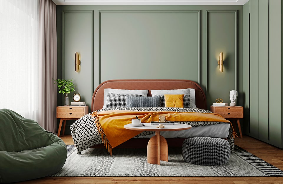 Bedroom in shades of dark green, natural wood and orange. The walls are dark green: the back one has frames and the side one houses a built-in wardrobe with doors in the same colour as the wall. The bed has a wooden frame, with a rounded-edged headboard. On either side are wooden bedside tables in modernist style, on which stand ornaments and vases. Above, fixed to the wall, are two upright golden lamps. The bed is on a black and white geometric-patterned carpet, and above the carpet are a pouffe and a small round wooden table with a book and a cup on it. On the left, next to the window, a large dark green ottoman can be seen;
