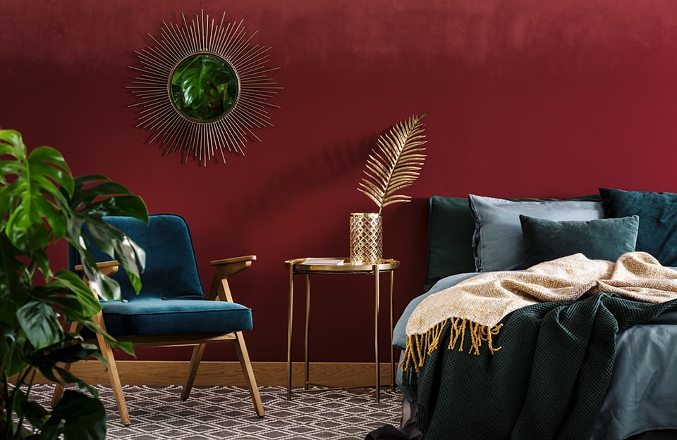 Detail of a bedroom with a red wall. The bed looks as if it has just been used and has dark linen in shades of grey-blue. Next to the bed is a small gold-plated metal table with a gold-plated basket on top and a gold-painted fern leaf inside. On the wall is a circular gilded sunburst mirror and underneath a Scandinavian-style armchair. To the left is a plant