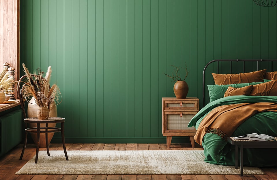 Bedroom in shades of green and natural wood. The wall is made of wood painted green. The floor is parquet. The bed, which is only partly visible, has a simple, light metal frame in black. Blankets, sheets and pillows are in shades of green and brown. At the side of the bed is an ethnic-style wooden bedside table with a vase with simple curved lines on top. On the floor is a beige carpet, with a chair on top of it that holds a wicker vase with dried herbs inside. Behind the chair is a large amphora-shaped vase. On the left is a window with a natural wood frame