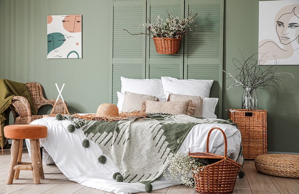Bedroom in modern rustic style, in shades of wood and green. Green wall, headboard of the bed in the same colour, made of old shutters. Parquet floor, geometrically patterned cotton blankets with ponpom and straw hat on the bed. In the room there are several wicker items (an armchair, two baskets, a bedside table with a vase on it, and many flowers and twigs. On the wall hang two pictures with stylised faces. At the end of the bed is a rustic stool with a pink seat;