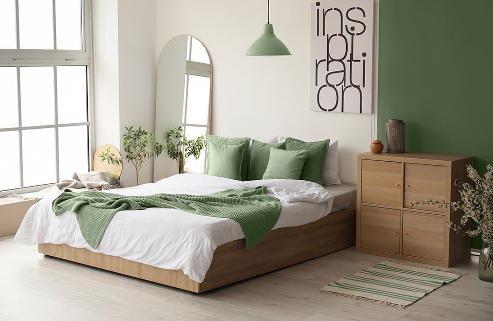 Modern bedroom in an essential style in a large room with a full-height window. The bed has a square and essential wooden frame, with green and white linen. The wall behind the bed is white and to one side, towards the window, is an arched mirror, behind a chair full of clothes. Above the bed is a graphic print with 'inspiration' written on it, and then a green wall begins, against which rests a square wooden cabinet with four doors, in front of which is a small carpet with geometric patterns. Above the bed, a green chandelier descends from the ceiling like the wall;