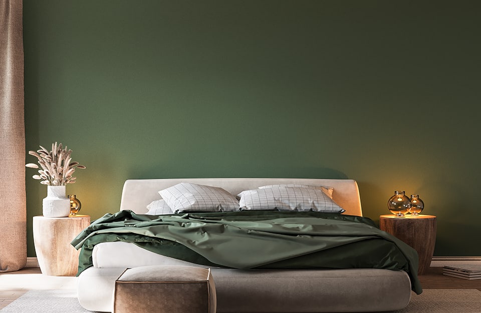 An elegant, minimalist bedroom with a green back wall, a large carpet under the bed and light coming in through a window on the left. The bed has a cream-coloured upholstered frame. The linen is white and green. On either side of the bed are two small round natural wood tables with vases on top;