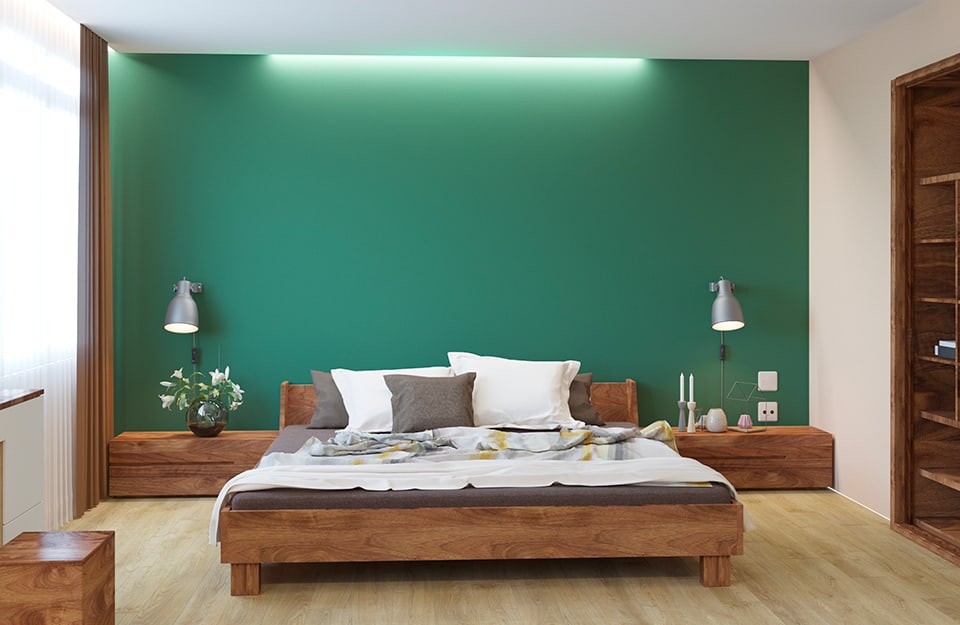 Bedroom with a green back wall illuminated by a strip of LEDs on the ceiling. The bed has a solid natural wooden frame, with a long, low wooden element behind it, running the length of the wall, which serves as a bedside table, with vases and candles resting on it. Two metal lamps hang on the wall on either side of the bed. The rest of the room is painted white. On the left side is a window, on the right a built-in wooden cupboard, open. The floor is light parquet;