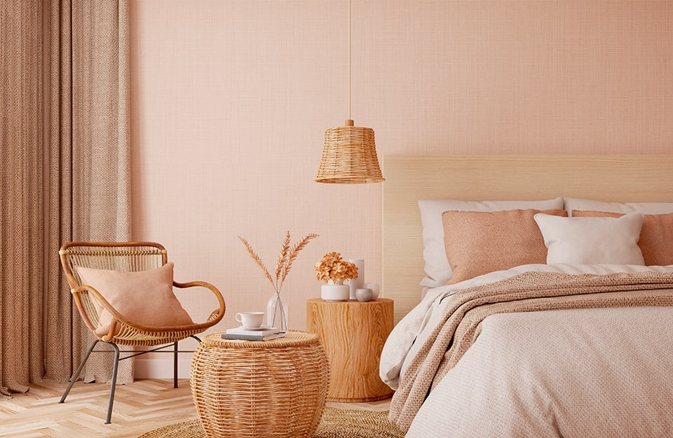 Bedroom in modern rustic style in beige and pastel tones. The bed has a simple light wooden frame. The linen is white and pastel pink. Next to the bed is a cylindrical bedside table made of natural wood with white jars on top. A bell-shaped wicker chandelier descends from above. Further to the left is a wicker coffee table with books, a vase and a cup on it, and a wooden armchair. The floor is light parquet and beige curtains can be seen;