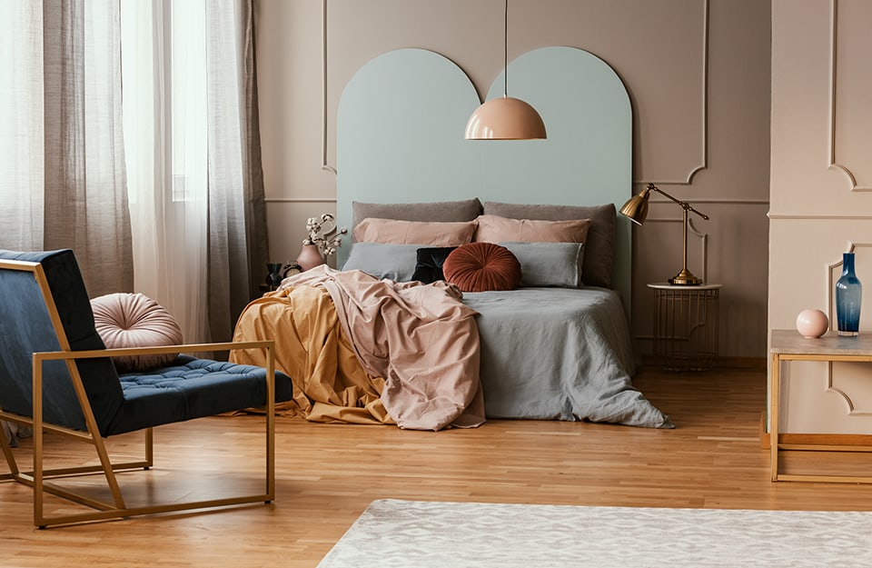 Bedroom in pastel tones, with beige walls decorated with frames. The bed has a very high pastel blue headboard ending in a double arch. A chandelier with a pastel-pink semicircular ceiling lamp descends from the ceiling. Sheets and pillows are also in various pastel tones. Next to the bed is a metal-framed bedside table with a lamp on top. At the end of the bed is a metal-framed armchair with a blue upholstered seat and back. There is also a white carpet on the floor and, leaning against a corner wall, another metal-framed, horizontal table with vases on top;