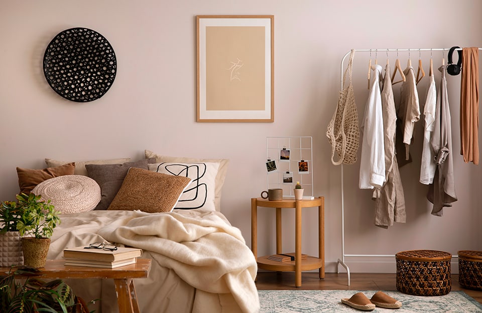 Bedroom in shades of beige, full of elements. The wall is beige, with just a minimal print and a wicker circle painted black. The bed is full of pillows and a blanket, in pastel shades of beige and brown. At the end of the bed is a spartan wooden bench, worn by time, with books, a pair of glasses and plants on it. To one side of the bed, a small round wooden table with a cup, a jar and a photo grid on top. Still to the right, a white metal drying rack with various clothes and accessories hanging from it. Below, a rug, wicker baskets and a pair of slippers;