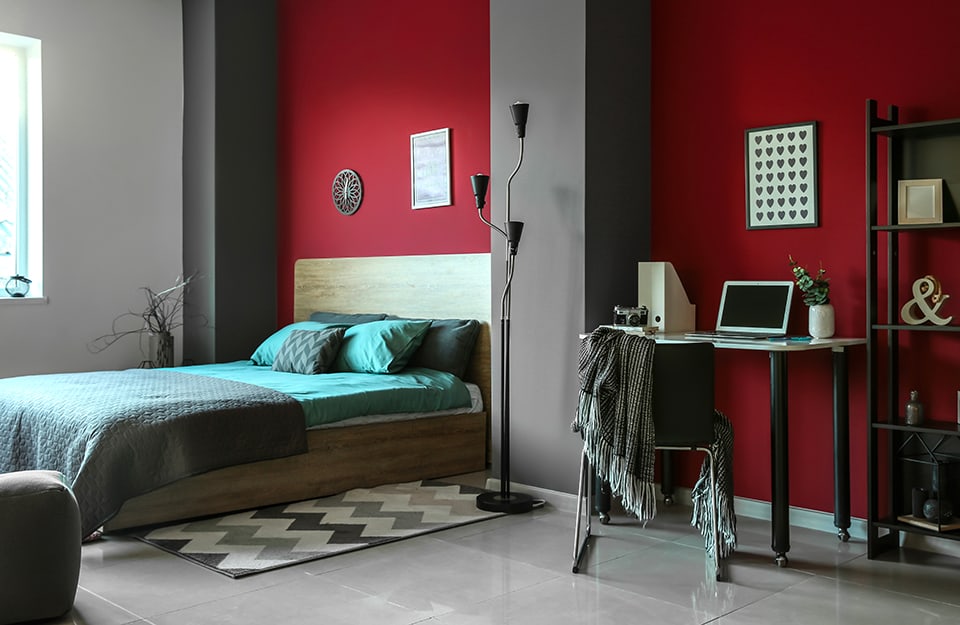 Bedroom with red and grey walls. The double bed has grey and blue linen. There is a desk with a chair and a bookcase. Two pictures on the walls and several knick-knacks