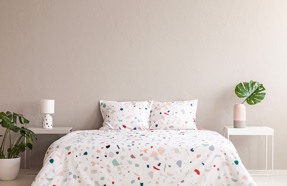 A minimal bedroom with a beige wall, a bed with a blanket and white pillows with colourful terrazzo-style elements, a motif that is also repeated on the bedside lamp, which is located on one of the two bedside tables, both in white metal and minimal. On the other bedside table is a simple two-tone grey and pink vase with a monstera leaf inside. A potted plant appears on the left