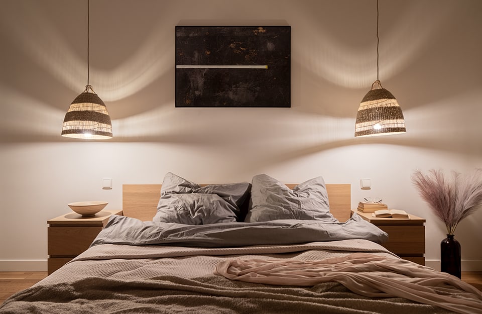 Bedroom in modern rustic style in the colours beige and brown. The wall is beige, with woven wooden lamps descending from the ceiling and casting undulating shadows. The bed has a simple, natural wooden frame. On either side of it are minimal wooden bedside tables with drawers and books and bowls on top. Next to one of the bedside tables is a bottle-shaped vase with plumes inside. Above the bed is a very dark abstract painting;