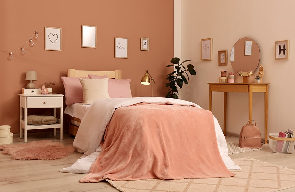 Bedroom in pastel shades of beige and powder. You can see one wall on hazelnut and one on beige, with many small pictures with writing and letters of the alphabet hanging on it. The bed is single, with a simple, wooden frame and many blankets in shades of powdery, pink and beige. Next to the bed is a rustic-style bedside table painted white, with objects on it and a lamp. On either side of the bed are three different rugs (one furry, and two short-haired). On the side of the bed opposite the bedside table is a plant. Leaning against the wall is a wooden dressing table with many objects on it and a circular mirror. On the floor is a bag and a magazine basket;